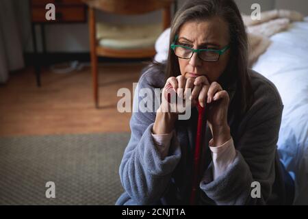 Preoccupata donna caucasica che passa il tempo a casa, la distanza sociale e l'isolamento in quarantena, seduta in camera da letto, tenendo il bastone da passeggio. Foto Stock