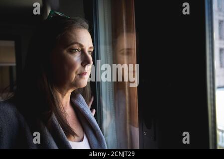 Donna caucasica con lunghi capelli scuri godendo del tempo a casa, distanziando sociale e auto isolamento in quarantena, in piedi e guardando fuori di wi Foto Stock