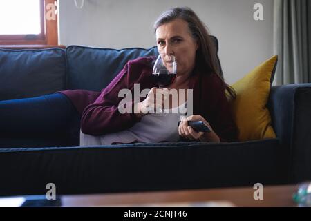 Donna caucasica che gode di tempo a casa, di distanza sociale e di isolamento in quarantena, sdraiata sul divano in salotto, bere vino rosso, Foto Stock