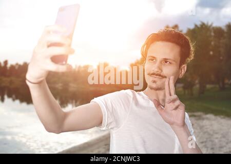 Ritratto di giovane bel ragazzo sorridente di stile fa selfie contro il lago. Segno di pace Foto Stock