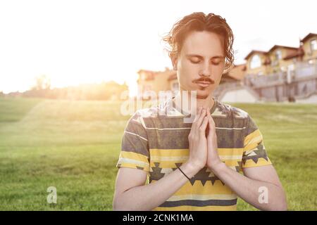 Giovane uomo meditando all'aria aperta nel parco, seduto con gli occhi chiusi e le mani insieme.godendo della natura, yoga e meditazione Foto Stock