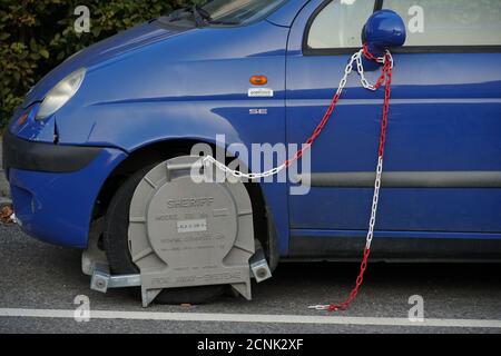Morsetto della ruota montato su una ruota di un veicolo parcheggiato in modo errato. Una catena rossa e bianca è fissata al morsetto e l'altra estremità allo specchietto laterale Foto Stock