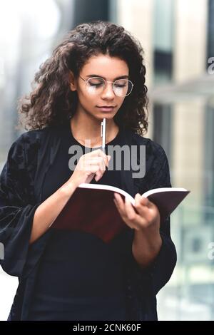 Ritratto di una donna di razza mista sorridente che prende appunti in un blocco note mentre si sta in piedi all'aperto. Foto Stock