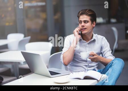 Giovane uomo sicuro con un abbigliamento casual elegante che tiene una tazza e parla con uno smartphone. Foto Stock