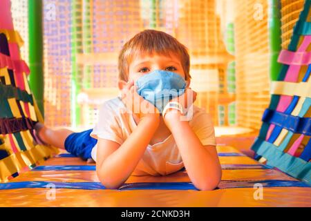 Scolaro in maschera protettiva giacente sul pavimento in sala giochi. Scuola elementare durante la quarantena. Ragazzo allegro in maschera in asilo. Sociale Foto Stock