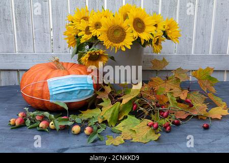 Un vaso di girasoli, una grande zucca su di esso, una maschera medica, foglie d'acero sono sul tavolo. Foto all'aperto. Concetto Proteggiti da pandemia, coronavirus. Foto di alta qualità Foto Stock