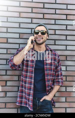 Parlare per telefono. Vista laterale del bel giovane uomo in abbigliamento casual elegante che parla dal telefono cellulare sullo sfondo della parete di mattoni. Foto Stock