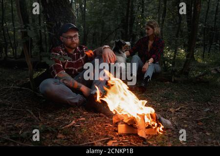 Bella Backpacker Donna e uomo che riposa vicino Bonfire dopo aver passeggiato ed escursionistico in foresta con Aussie Australian Breed Dog. Contiene crominanc Foto Stock