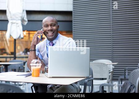Un imprenditore nero molto sorridente che parla al telefono e lavora a distanza con il suo laptop. È seduto sulla terrazza di una caffetteria. Spazio per Foto Stock