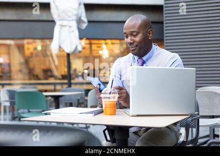 Uomo d'affari afro-americano che usa il suo smartphone mentre lavora a distanza da una moderna caffetteria. Spazio per il testo. Foto Stock