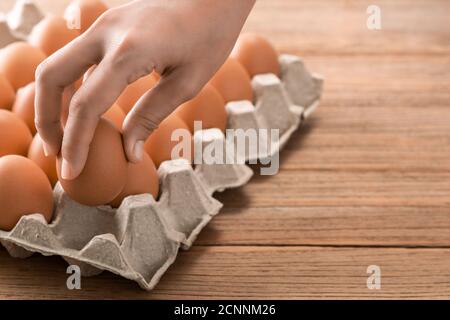 Mano delle donne che prendono uovo di gallina marrone da molte uova fresche di pollo marrone crudo in scatola di cartone. Vintage tavolo da cucina in legno sfondo. Foto Stock