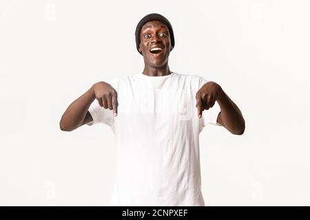 Ritratto di un bel maschio afro-americano eccitato e ottimista, puntando le dita verso il basso e sorridendo stupito, sfondo bianco Foto Stock