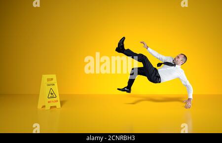 businessman scivola sul pavimento bagnato, segno di attenzione in vista, sfondo giallo. Foto Stock