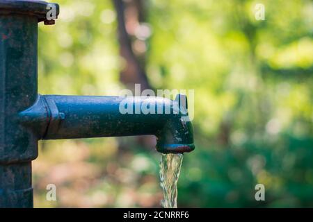 Pompa manuale a leva verde e arrugginita con acqua versando fuori dal beccuccio con uno sfondo verde sfocato Foto Stock