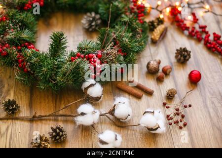 Decorazioni natalizie e corona natalizia con luci dorate su sfondo rustico in legno. Cotone, bacche rosse, ornamenti e coni di pino per la tradizione Foto Stock