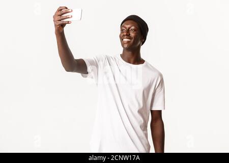 Ritratto di bel ragazzo afro-americano alto in bania, prendendo selfie su smartphone e sorridente felice, sfondo bianco Foto Stock