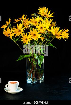 Bouquet di fiori gialli freschi luminosi e tazza bianca di caffè su sfondo nero. Fiori di carciofo di Gerusalemme in vaso trasparente di vetro. Tasto basso Foto Stock