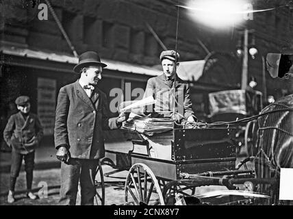 Immagine di un newsboy con una disabilità in un carrello trainato da cavalli sul lato di una strada a Chicago, Illinois, che consegna un giornale a un uomo. Foto Stock