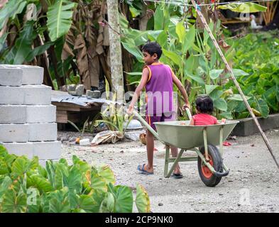 FUAHMULAH ISLAND, MALDIVE - 11 APRILE 2017: Un ragazzo rotola la sorella in una carriola mentre i loro genitori costruiscono una casa Foto Stock