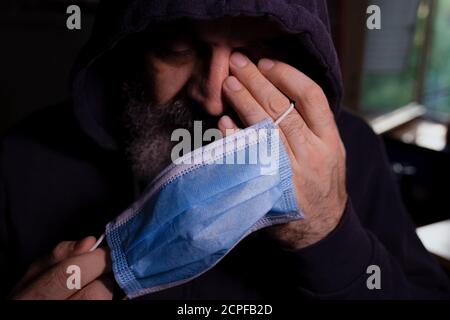 Uomo bearded in hoodie scuro che tiene la maschera chirurgica blu, gli occhi chiusi, una mano sul viso, stanco e depresso guardare Foto Stock
