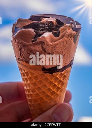 Cono gelato al cioccolato tenuto contro un cielo estivo con nuvole e sole con raggi nell'angolo in alto a destra Foto Stock