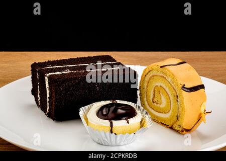 Tre tipi di dessert, rotolo di torta, cupcake e torta al burro di cioccolato in piastra di ceramica bianca su tavolo di legno. Foto Stock