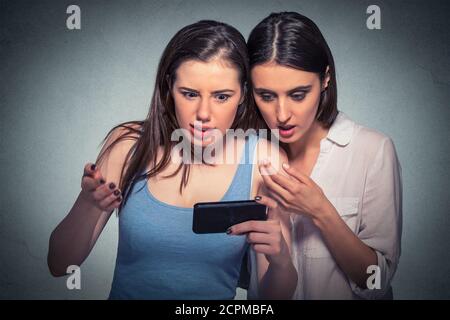 Due ragazze sorprese che guardano il telefono delle cellule che discutono le ultime notizie del gossip, fiabbergasted a che cosa vedono lo sfondo grigio isolato Foto Stock
