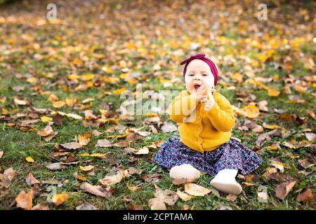 Baby girl in giacca gialla e archetto rosso seduta sull'erba, giocando nelle foglie autunnali. Foto Stock