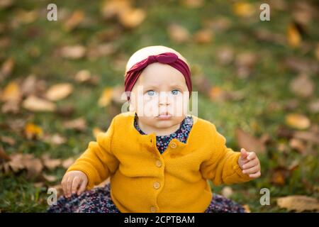 Baby girl in giacca gialla e archetto rosso seduta sull'erba, giocando nelle foglie autunnali. Foto Stock