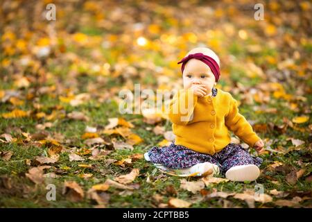 Baby girl in giacca gialla e archetto rosso seduta sull'erba, giocando nelle foglie autunnali. Foto Stock