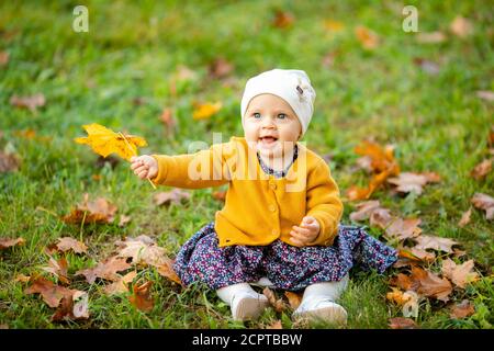 Baby girl in giacca gialla e archetto rosso seduta sull'erba, giocando nelle foglie autunnali. Foto Stock