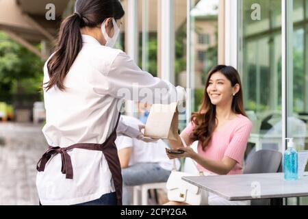 Vista posteriore della cameriera asiatica con maschera faccia dare ordine di portare fuori sacchetto di cibo a donna attraente cliente femminile. Servizio di ristorazione da take-away o da take-out con Foto Stock