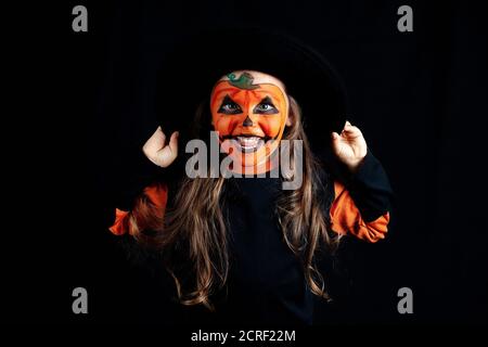 Ragazza con i capelli sciolti con il trucco della zucca che tiene le mani sul bordo di un cappello nero e sorridente, Halloween Foto Stock