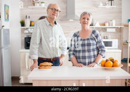 Ritratto della vecchia coppia allegra che guarda in macchina fotografica in cucina durante la preparazione della colazione. Felice uomo e donna in pensione. Foto Stock