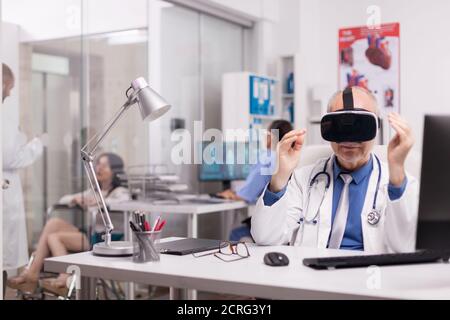 Medico senior che indossa cuffie per la realtà virtuale in ufficio ospedaliero, mentre un giovane terapeuta discute con una donna disabile in sedia a rotelle e un infermiere in uniforme blu. Foto Stock