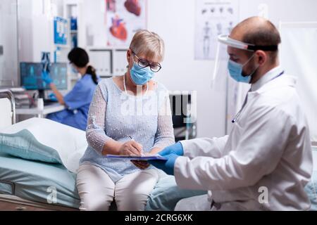 Donna anziana che firma i risultati del test medico durante la consultazione con il medico nella sala di esame dell'ospedale e che indossa la maschera facciale agasint covid per la precauzione di sicurezza. Foto Stock