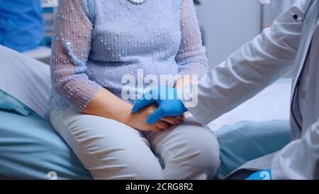 Primo piano di mani medico in blu guanti protettivi comfortin una donna anziana stressata seduta sul letto dell'ospedale in clinica privata moderna. Pandemia globale coronavirus covid-19 pandemia sanitaria Foto Stock