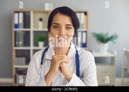 Giovane medico amichevole che guarda la macchina fotografica durante la consultazione in linea dentro il suo moderno ufficio ospedaliero Foto Stock