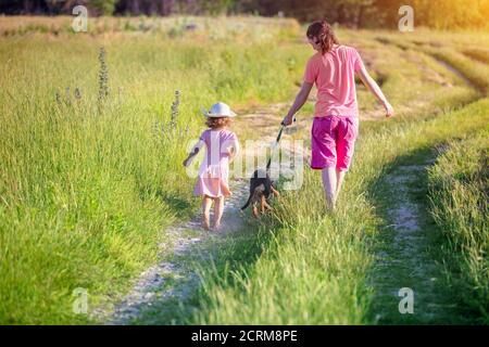 Madre felice che cammina con una piccola figlia a piedi nudi all'aperto in estate. La madre conduce un cane sul guinzaglio ang passeggiate sulla strada sterrata paese. Felice Foto Stock