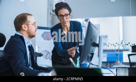 Modern Factory: La Supervisore del progetto femminile parla con un ingegnere industriale maschile che lavora su Personal computer. Utilizzano il software CAD per la progettazione e. Foto Stock