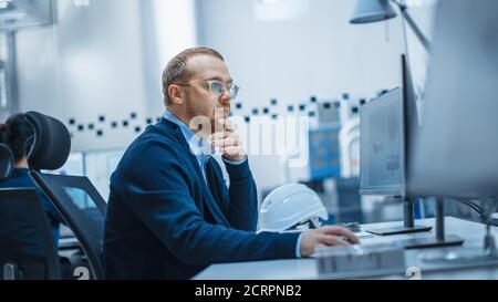 Colpo di Male Industrial Engineer risolvere i problemi, lavorando su un Personal computer, sta lavorando in Ufficio in fabbrica moderna con High-Tech Machinery. Foto Stock