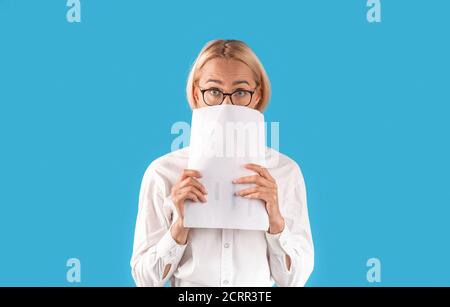 Scioccato imprenditore femminile peeking da dietro documenti su sfondo blu studio, panorama Foto Stock