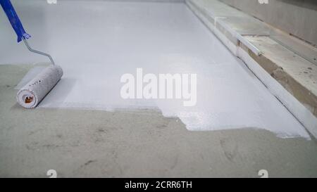Un lavoratore dipinge il pavimento in cemento con un rullo bianco. Lavori di riparazione del pavimento in cemento. Rullo di vernice in cantiere Foto Stock