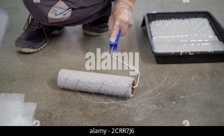 Un lavoratore dipinge il pavimento in cemento con un rullo bianco. Lavori di riparazione del pavimento in cemento. Rullo di vernice in cantiere Foto Stock