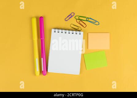 Vista dall'alto del notebook vuoto e dei materiali di consumo per la scuola, come pennarelli colorati, adesivi e fermi su sfondo giallo, spazio per il testo. Foto Stock