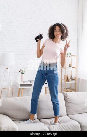 Musica preferita a casa durante l'isolamento. Donna afro-americana felice in cuffia danze sul divano Foto Stock