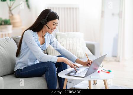 Apprendimento a distanza. Bella donna asiatica in cuffia studiare con il portatile a casa Foto Stock