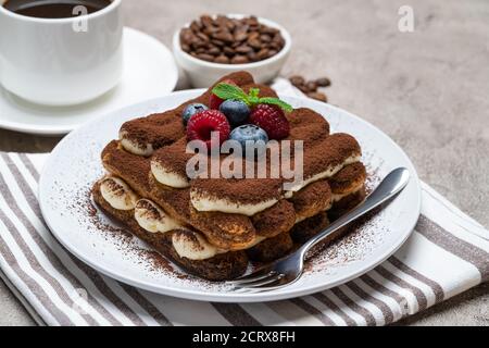 Porzione di dessert classico tiramisù con lamponi e mirtilli, tazza di caffè e panna o latte su fondo grigio di cemento Foto Stock