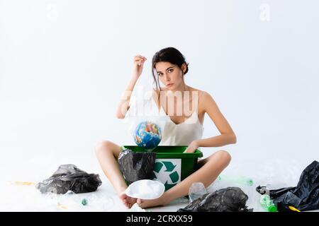 giovane donna che tiene sacchetto di plastica con il globo vicino ai rifiuti e. cestino con simbolo di riciclaggio su bianco Foto Stock