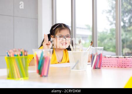 Ragazza asiatica felice con la sindrome di Down alzare due dita e sedersi alla scrivania piena di strumenti d'arte. Foto Stock